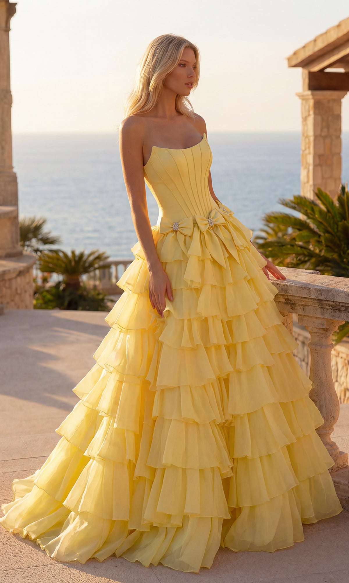 A woman in a light yellow Tiered Prom Ball Gown with Bows (PromGirl 250071) stands outdoors on a terrace, with the ocean and palm trees in the background.